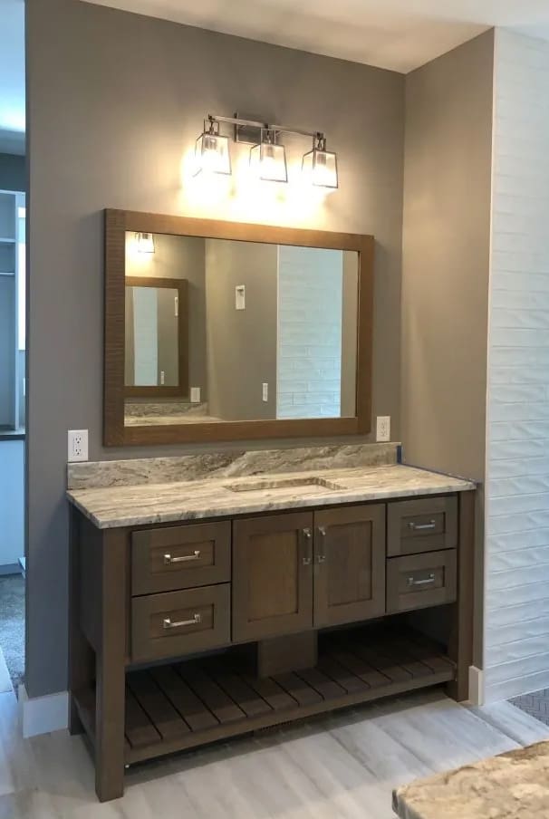 A modern bathroom vanity with a wooden cabinet, marble countertop, rectangular mirror, and three-light fixture above. The vanity has drawers, cabinet doors, and an open shelf underneath. Walls are neutral and floor is light-colored tile.