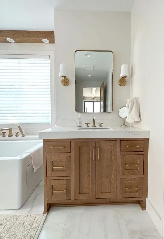 A modern bathroom features a wooden vanity with gold hardware, a white countertop, a rectangular mirror with gold trim, two wall sconces, and a bathtub next to a window with white blinds.
