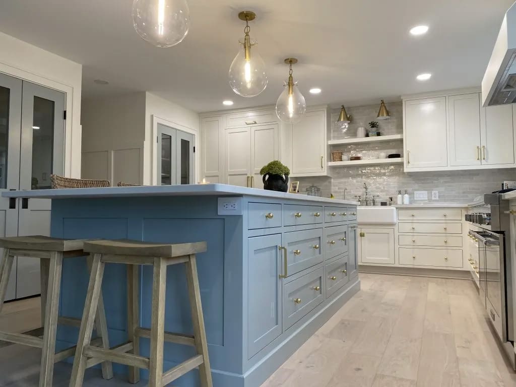 Modern kitchen with a large blue island, wooden stools, white cabinets with gold handles, farmhouse sink, stainless steel appliances, pendant lights, open shelves, and light wood flooring.