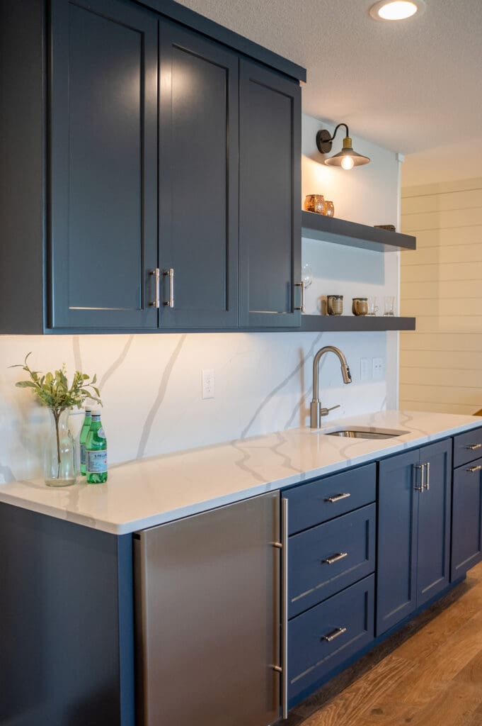 Modern kitchen with navy blue cabinets, white marble-patterned countertops and backsplash, stainless steel sink and faucet, mini fridge, open shelves with glassware, and a wall sconce above the shelves.