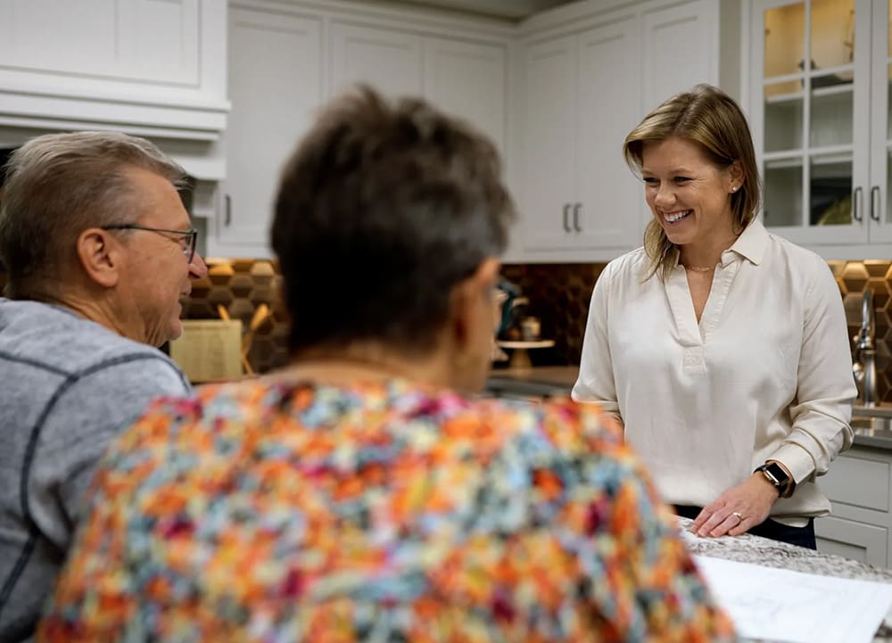 A woman standing and smiling talks with two seated people in a modern kitchen with white cabinets and a brown hexagonal tile backsplash.