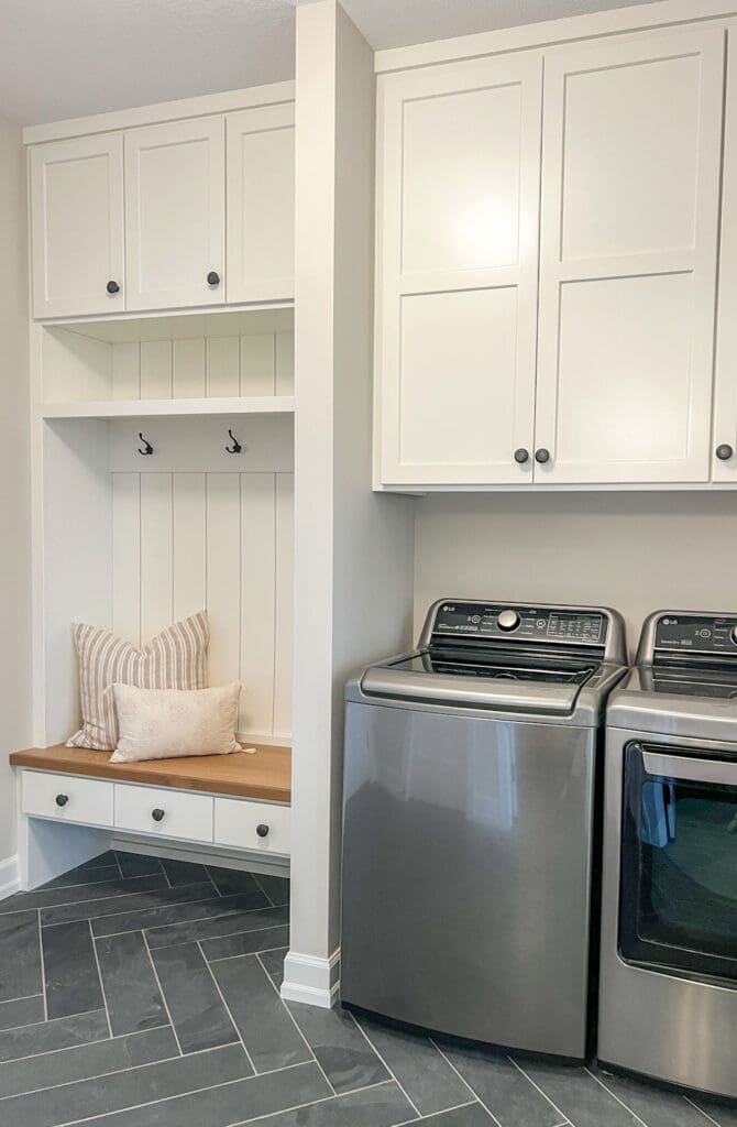 A modern laundry room with white cabinets, a stainless steel washer and dryer, a built-in bench with a striped pillow, three coat hooks, and herringbone-patterned tile flooring.