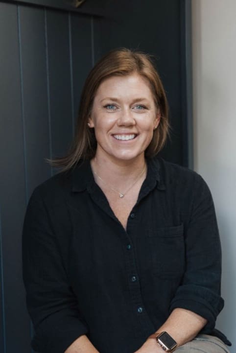 A woman with shoulder-length brown hair and blue eyes smiles at the camera. She is wearing a black button-up shirt, a necklace, and a smartwatch, sitting in front of a dark background.