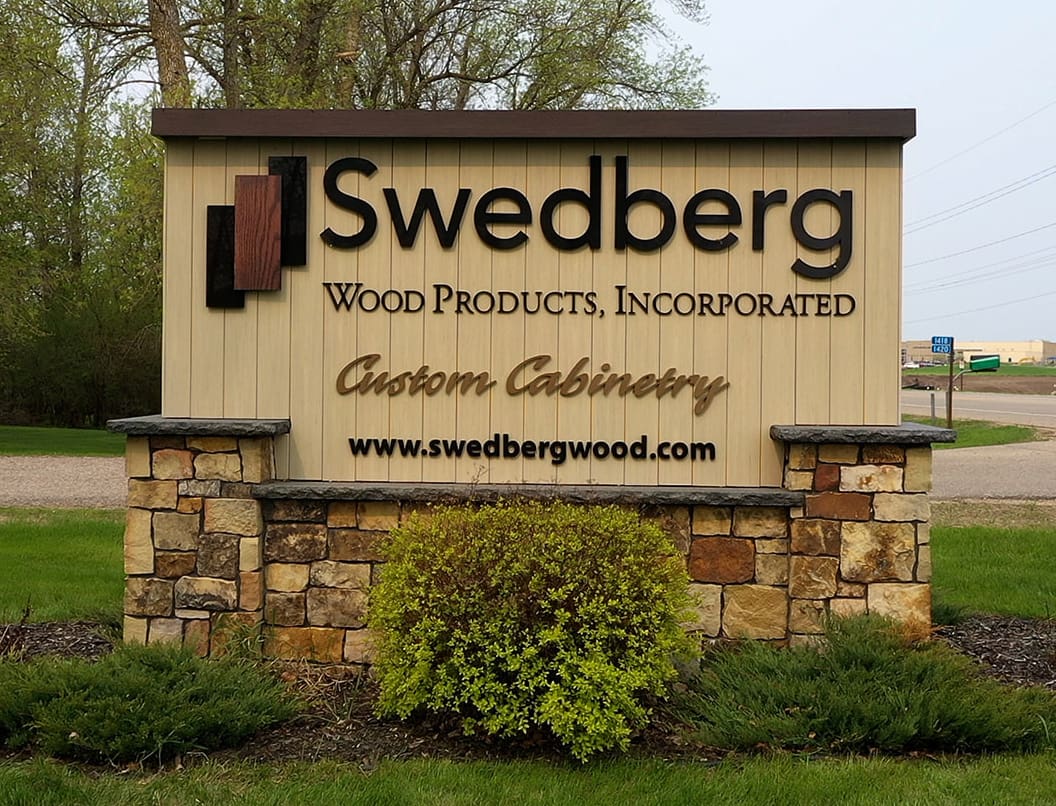 Outdoor sign for Swedberg Wood Products, Incorporated, advertising custom cabinetry with their website, www.swedbergwood.com, on a beige and stone structure with bushes and grass in front.
