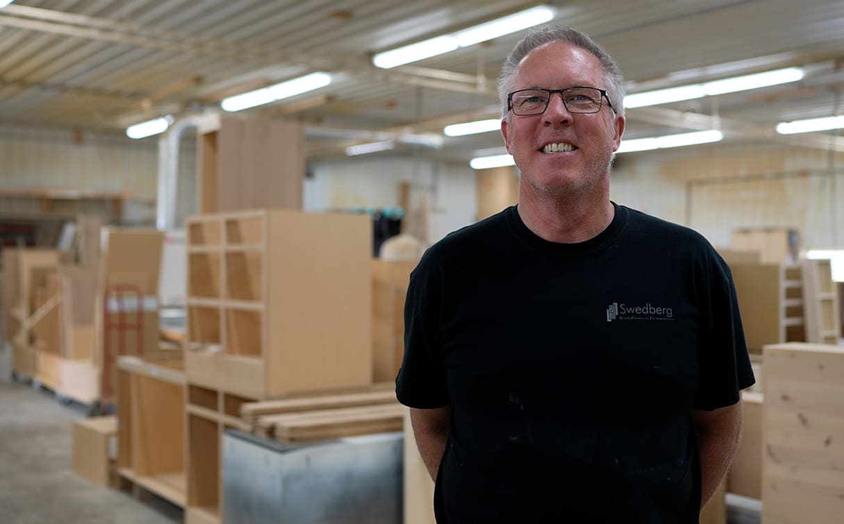 A smiling man wearing glasses and a black "Swedberg" t-shirt stands in a well-lit woodworking shop, surrounded by unfinished wooden cabinets and materials.