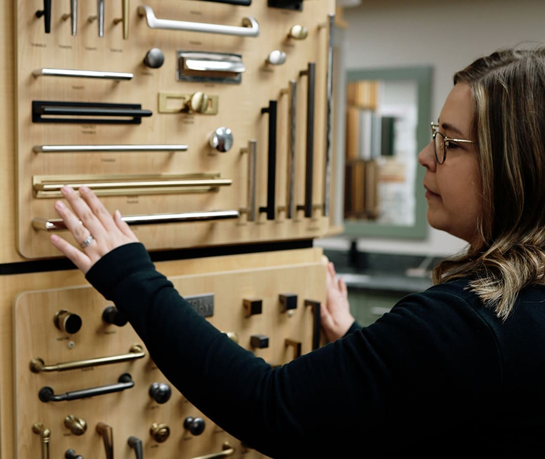A woman wearing glasses examines a display board with various styles and sizes of handles and knobs, possibly in a hardware or home improvement store.