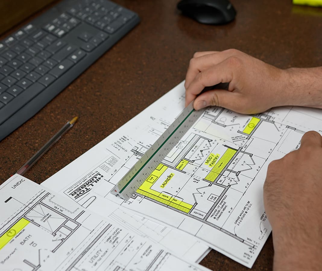 A person uses a ruler to measure and review architectural floor plans on a desk, with a keyboard, mouse, and pencil nearby. Some sections on the plans are highlighted in yellow.