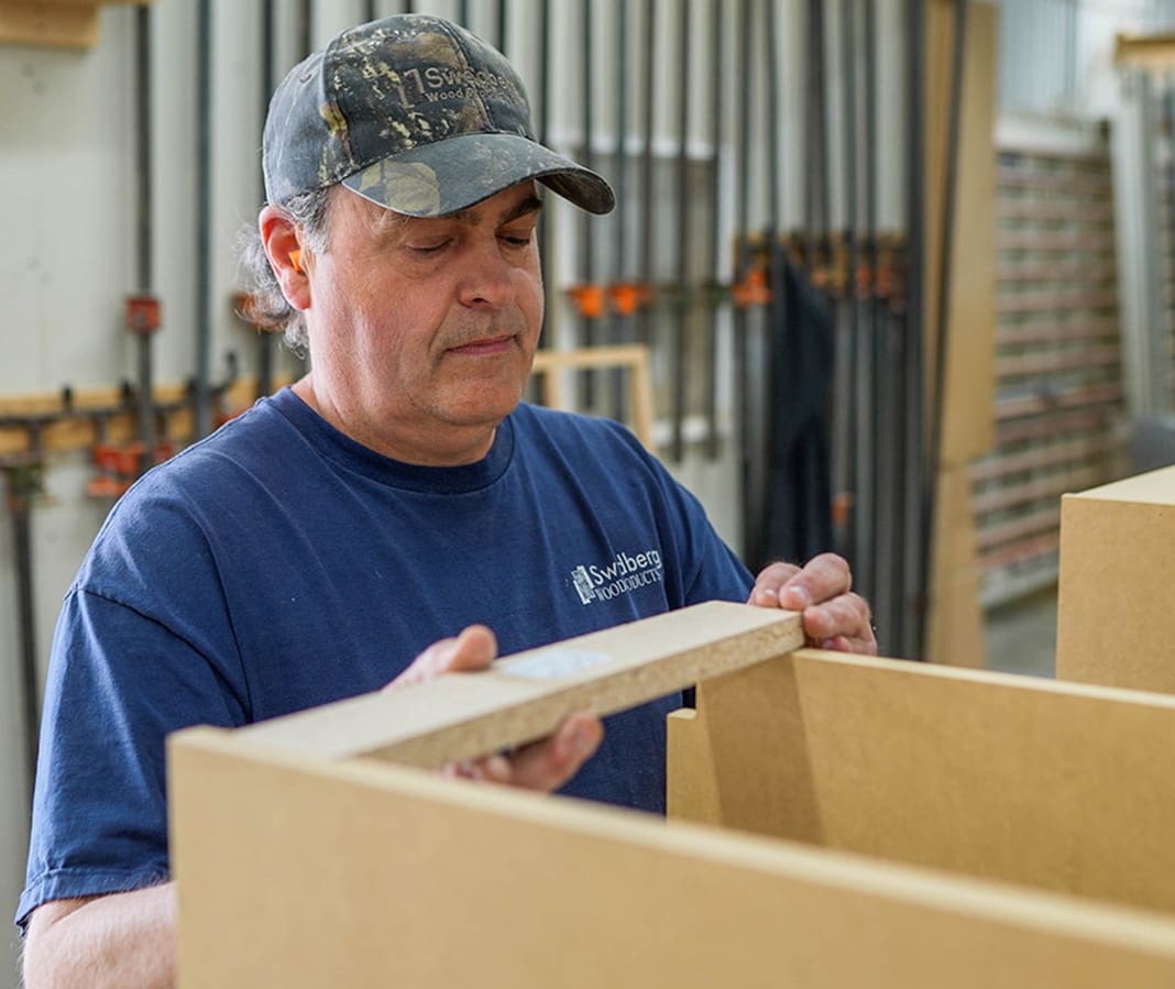 A man wearing a camouflage cap and blue t-shirt carefully measures a wooden board in a workshop, surrounded by woodworking tools and materials.