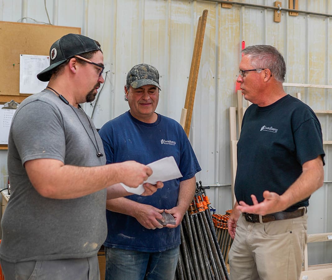 Three men stand in a workshop having a discussion. One man holds papers, another is smiling, and the third gestures with his hands. Tools and wooden boards are visible in the background.