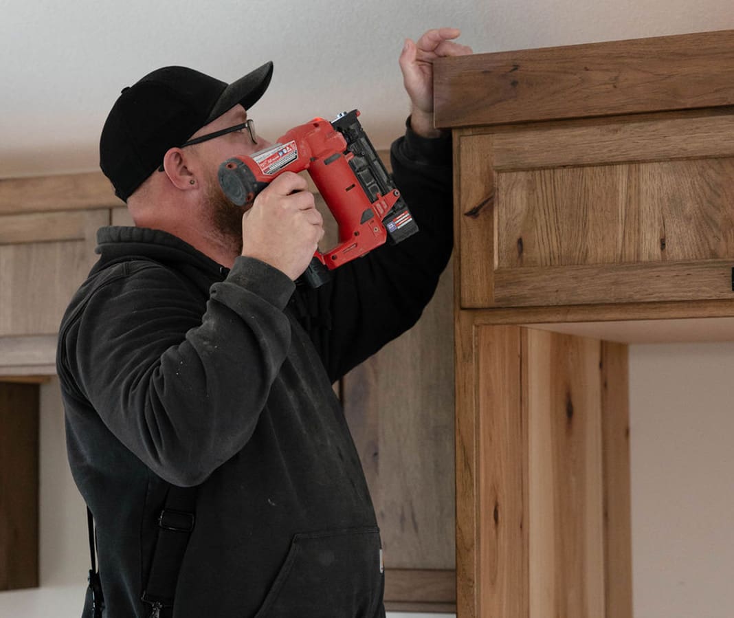 A person wearing glasses, a black cap, and a dark hoodie uses a red nail gun to fasten wood trim above a set of wooden cabinets inside a home.