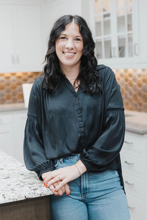 A woman with wavy dark hair, wearing a black blouse and blue jeans, smiles while leaning on a granite kitchen counter. The kitchen has white cabinets, glass doors, and a brown hexagonal tile backsplash.