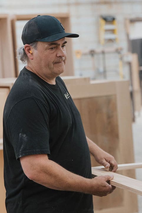 A man wearing a black t-shirt and black cap stands in a workshop, holding a piece of wood. The background shows wooden furniture pieces and workshop equipment.