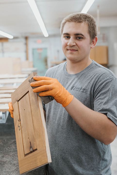 A young man wearing orange gloves and a gray T-shirt sands a wooden cabinet door in a workshop, looking at the camera with a slight smile. Bright lights and woodworking tools are visible in the background.