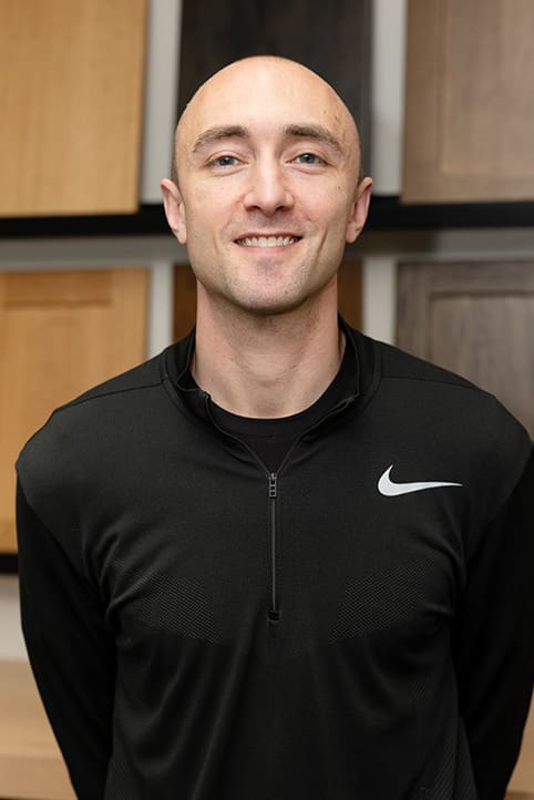 A man with a shaved head smiles at the camera. He is wearing a black Nike zip-up jacket and standing indoors in front of wooden panels in various colors.