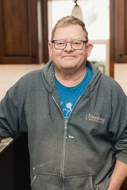 A middle-aged man with glasses wears a gray zip-up hoodie and a blue T-shirt, standing indoors in a kitchen with brown cabinets, smiling gently at the camera.