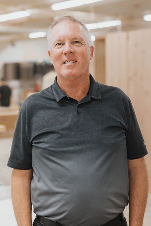 A middle-aged man with short gray hair, wearing a dark gray polo shirt, stands indoors in a workspace with wooden surfaces and blurred people in the background.