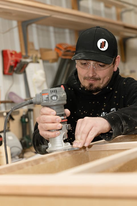 A person wearing safety glasses and a black cap uses a handheld woodworking router on a piece of wood, with sawdust flying in a workshop.