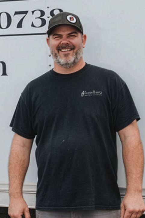 A smiling man with a gray beard and mustache wears a black cap and black T-shirt while standing outdoors in front of a white vehicle.