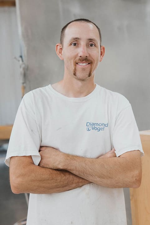 A man with short brown hair and a mustache stands with arms crossed, smiling. He wears a white "Diamond Vogel" T-shirt and is in an indoor workspace with industrial equipment in the background.