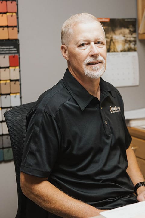A middle-aged man with short light hair and a goatee sits at a desk, wearing a black polo shirt. Behind him are paint swatches on the wall and a calendar. He looks at the camera with a neutral expression.