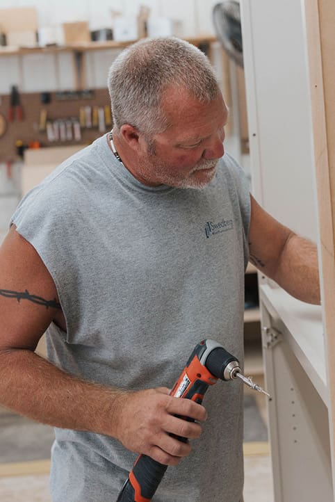 A man with short gray hair and a beard, wearing a sleeveless gray shirt, uses an electric drill while working on a wooden project in a workshop.