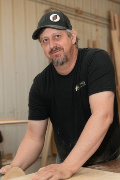 A man wearing a black cap and T-shirt is smiling while working with a piece of wood in a workshop, surrounded by wooden planks and tools.