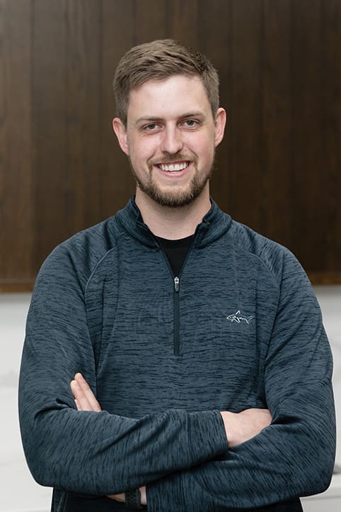 A man with short brown hair and a beard smiles with his arms crossed, wearing a dark quarter-zip pullover. The background features a wooden panel above a white wall.