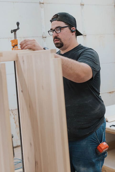 A man wearing glasses, a backwards cap, and earphones works on a wooden cabinet using a clamp in a workshop.
