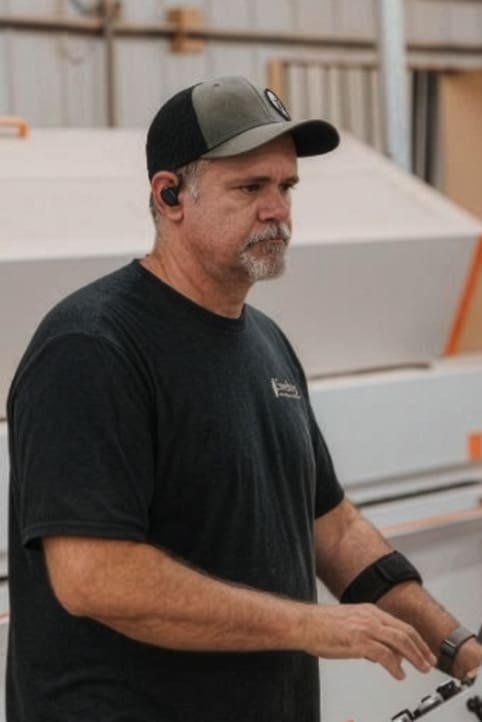A middle-aged man with a gray beard wears a black T-shirt, a black and gray cap, and wireless earbuds, standing indoors near industrial equipment.