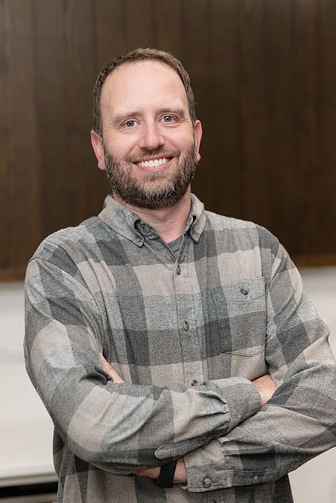 A man with a beard and short hair smiles with arms crossed, wearing a gray and black plaid shirt. The background features a dark wooden wall.