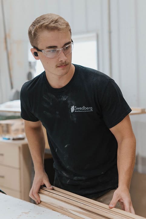 A young man wearing safety glasses and a black “Swedberg” T-shirt stands in a workshop holding wooden boards, appearing focused on his woodworking project.