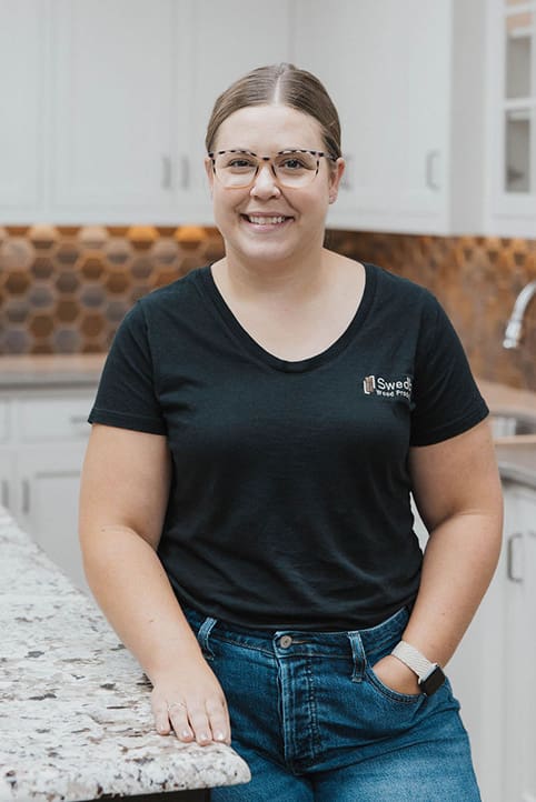 A woman with glasses, wearing a black t-shirt and blue jeans, smiles while standing in a modern kitchen with white cabinets and a hexagon-tiled backsplash.