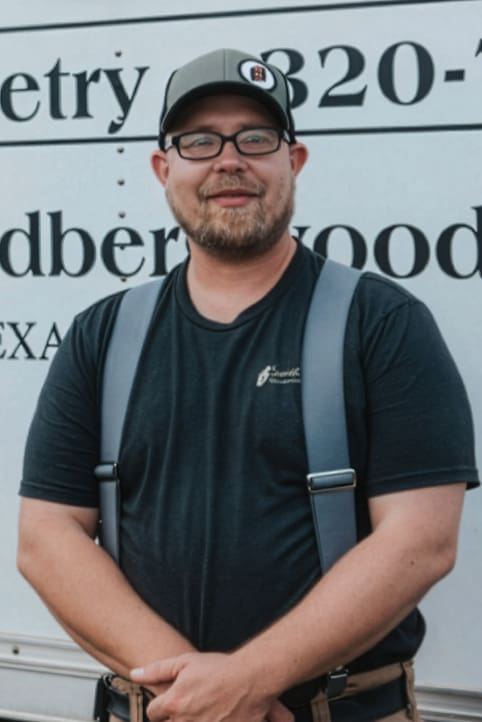 A man with glasses and a beard, wearing a black T-shirt, suspenders, and a cap, stands smiling in front of a white truck with partial text visible in the background.