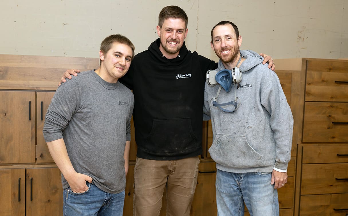Three men stand smiling together in a workshop, wearing casual clothes and hoodies. One man has his arm around the others, and another has a respirator around his neck. Wooden cabinets are in the background.