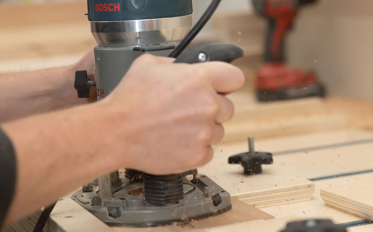 Close-up of a person using a Bosch electric router to shape or carve wood, with sawdust flying and other tools blurred in the background.
