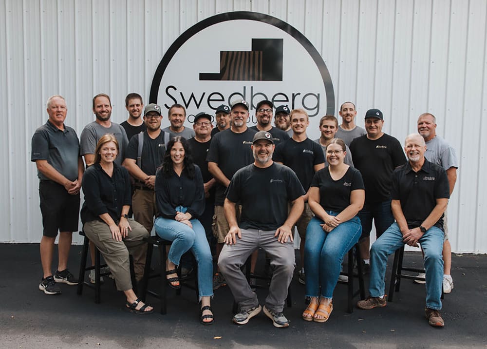 A group of 19 people, men and women, pose and smile in front of a white building with a "Swedberg Wood Products" logo on the wall. Some are seated in front while others stand behind them.
