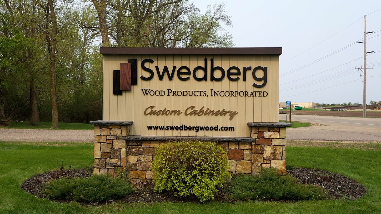 A sign for Swedberg Wood Products, Incorporated, advertising custom cabinetry and displaying their website, swedbergwood.com, stands on a stone base with grass and trees in the background.